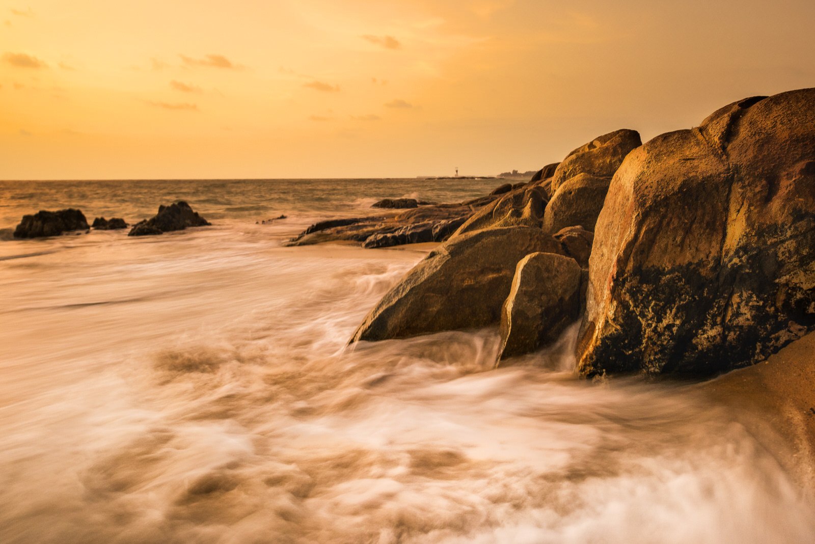 Waves crash against large rocks on a coastline under an orange-yellow sky at sunset or sunrise.
