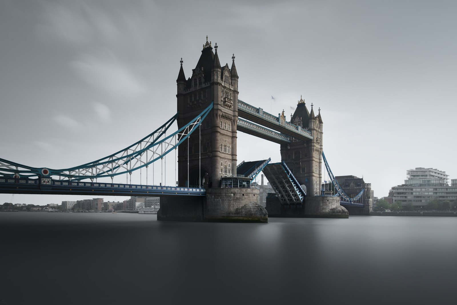 Tower Bridge in London, with its bascules partially raised, spans the River Thames under an overcast sky.