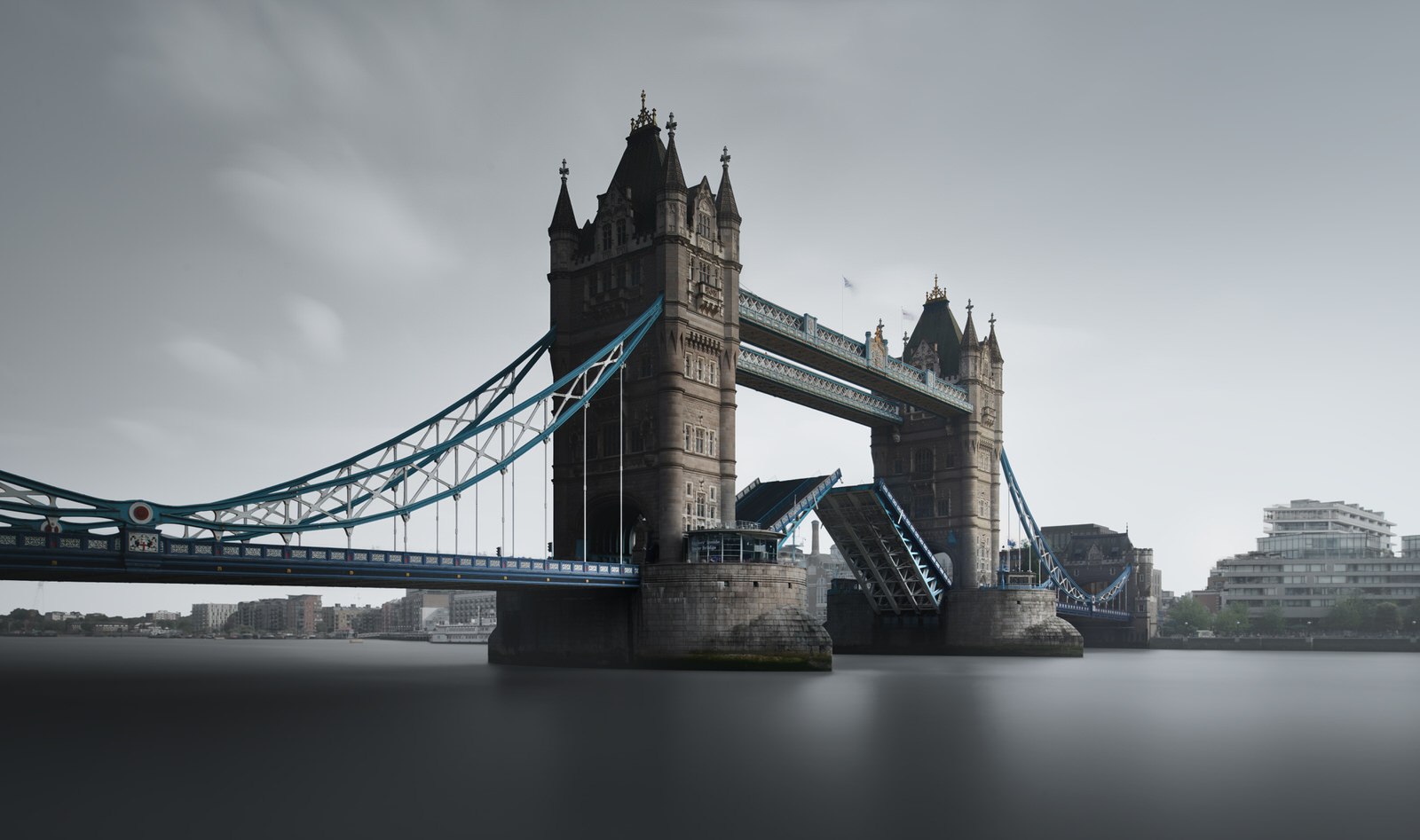 Tower Bridge in London, with its bascules partially raised, spans the River Thames under an overcast sky.