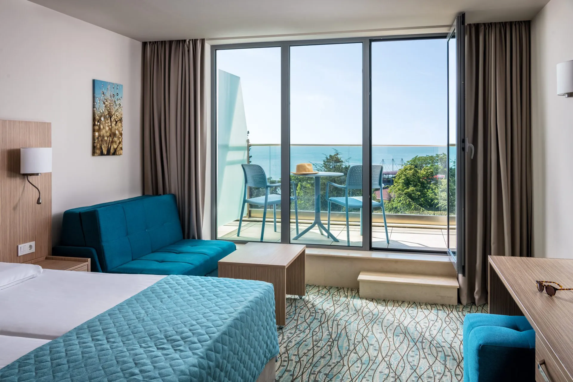 Bright hotel room with floor-to-ceiling glass doors, ocean view, blue accent sofa, and private balcony